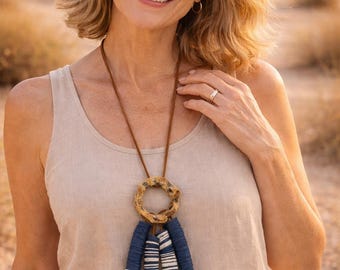 May include: A woman wearing a tan vest top and a brown leather necklace. The necklace features a circular, textured pendant and three blue and white beaded tassels. The background is a blurred desert landscape.