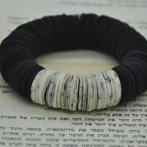 Op de afbeelding: Een zwart-witte cirkelvormige armband. De armband is gemaakt van gestapeld, afgerond zwart materiaal met secties van wit, getextureerd materiaal. De armband ligt op een pagina met tekst.
