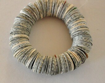 May include: A circular bracelet constructed from numerous small, rectangular paper pieces. The paper displays printed text, with off-white and grey edges. The bracelet is presented on a white surface.