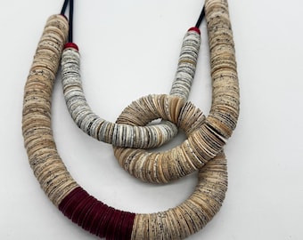 Puede incluir: Un collar de varias capas con numerosas cuentas pequeñas, circulares y similares al papel en tonos beige y gris con patrones moteados. El diseño incluye una sección de color rojo oscuro y pequeños acentos rojos en el cordón negro.