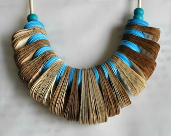 Puede incluir: Un collar hecho de páginas dobladas de un libro antiguo, con cuentas y cuerda azul turquesa. Las páginas están superpuestas y en abanico para crear un aspecto único y texturizado.