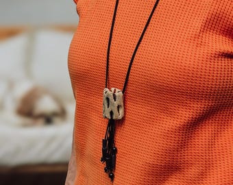 May include: A distinctive necklace with a light-coloured, textured pendant, a dark cord, and black beaded details. The pendant is set against an orange waffle-knit top and blue jeans.