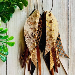 May include: Dangle earrings featuring layered leather strips in metallic gold, black, and brown with snakeskin and floral patterns. The earrings hang from bronze-toned circular hoops and include a delicate gold-toned chain with small round beads.