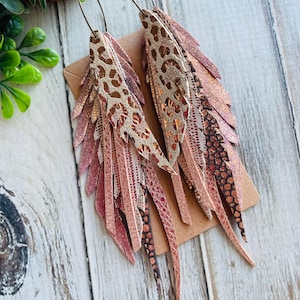 Peut inclure: Une paire de longues boucles d'oreilles en cuir rose et marron avec un design superposé et frangé. Les boucles d'oreilles sont composées de plusieurs morceaux de cuir superposés et frangés pour créer un effet de plume. Les boucles d'oreilles sont un bijou magnifique et unique qui ajouterait une touche de style à n'importe quelle tenue.
