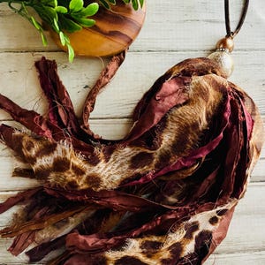 May include: A handmade tassel pendant featuring a mix of fabrics in burgundy, brown, and animal print patterns. The pendant is suspended from a brown cord and includes a decorative bead. The tassel is a unique accessory.