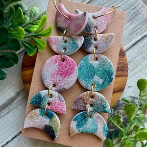 May include: A set of six handmade earrings with a glittery finish. The earrings feature a mix of pink, blue, and white colors. Each earring has a gold-colored hoop or connector. The earrings are displayed on a brown card.