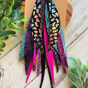 May include: A pair of dangle earrings with black, pink, and teal leather fringe. The fringe is iridescent and has a textured pattern. The earrings are attached to silver hoop earrings.