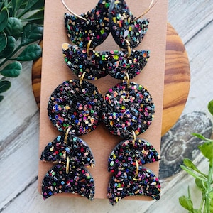 May include: Black dangle earrings with gold hoops and connectors. The earrings feature a layered design with a glittery, multi-colored confetti pattern. The earrings are displayed on a brown card.