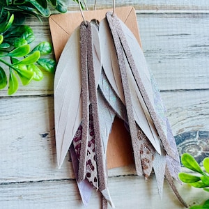 May include: A pair of dangle earrings with multiple layers of leather in shades of gray, white, and purple. The leather is textured and has a subtle shimmer. The earrings are attached to silver hoops.