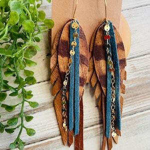 May include: A pair of handcrafted earrings featuring a feather design. The earrings combine brown leather, teal suede, and gold-colored chain accents. The feather-shaped earrings are attached to gold-colored ear wires.