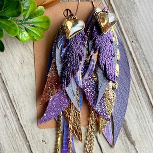 May include: A pair of dangle earrings featuring layered feather-shaped cutouts in purple, gold, and silver textures. Each earring is topped with a gold-tone heart-shaped crystal charm and includes dangling gold chains and rhinestone accents.