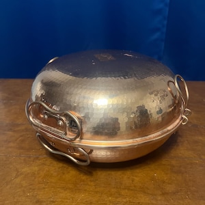 May include: A round, hammered copper cooking vessel with a lid, featuring two side handles and a metal latch mechanism. A small rectangular label on the side displays the text "METALUR" and "MADE IN PORTUGAL". The surface has a reflective finish.