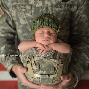 May include: A baby wearing a green knitted hat with buttons is being held by a person wearing a camouflage uniform with the words "U.S. ARMY" and "PIERCE" on it. The background is an American flag.