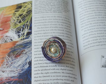 May include: A colourful, circular, paper-based brooch with a pearl-like button in the centre. The brooch is made of multiple layers of paper, each with a different pattern and colour. The brooch is lying on a page of a book.