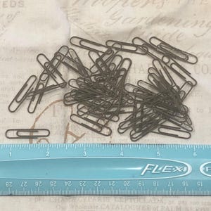 May include: A collection of bronze-colored paper clips scattered on a vintage paper background. A blue ruler with inch measurements is at the bottom of the image, providing scale. The paper clips are a classic office supply.