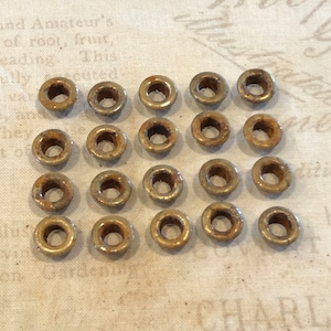 May include: A collection of twenty small, aged metal eyelets. Each eyelet has a circular shape with a central hole and a slightly rusted appearance. The eyelets are arranged in a grid pattern on a textured, off-white fabric.