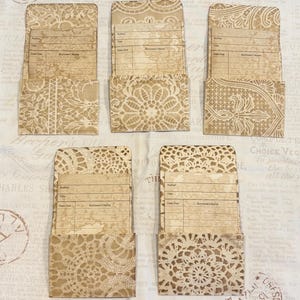 May include: Five rectangular library card pockets featuring a tan and cream lace pattern. Each pocket contains a paper card with printed fields for Author, Title, Date Due, and Borrower's Name. The paper has a vintage, aged, and stained appearance.
