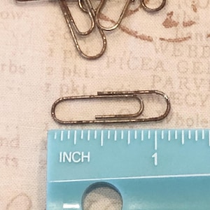 Könnte beinhalten: Nahaufnahme eines rostigen Büroklammern, der 2,5 cm lang ist. Die Büroklammer liegt auf einer weißen Stoffoberfläche neben einem blauen Lineal.