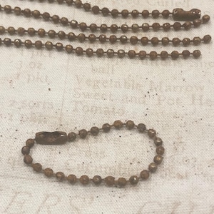 May include: Close-up of several antique bronze ball chains, some in straight lines and one looped into a circle. The chains have a textured, aged appearance, with a tarnished bronze color. The background is a light, textured fabric.