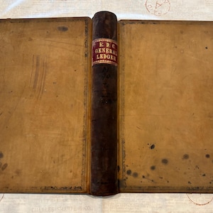 May include: An antique ledger book with a dark brown leather spine and tan covers. The spine features a red label with gold text reading R. D. C. GENERAL LEDGER. The covers have decorative border detailing and show signs of age with dark spots.