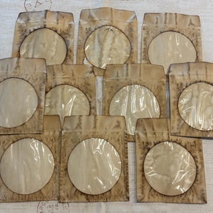 May include: A collection of ten rectangular paper envelopes featuring a distressed, tea-stained appearance with dark brown edges. Each envelope has a large, circular transparent plastic window centered on the front, allowing for visibility of the contents.