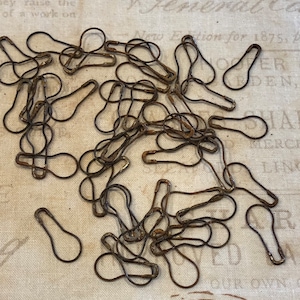 May include: A pile of antique safety pins, also known as dressmaker pins, in a rusty brown color. The pins are scattered on a white fabric surface with printed text.