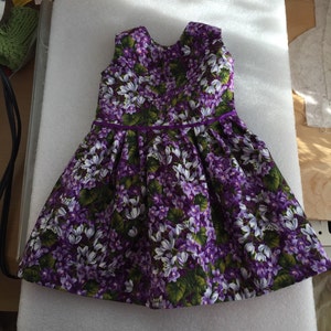 May include: A sleeveless dress featuring a floral pattern of purple and white flowers with green leaves. The dress has a pleated skirt and a purple ribbon detail at the waist. The dress is made of a lightweight fabric.