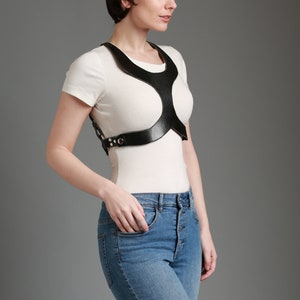 May include: A white t-shirt with a black leather harness. The harness has a unique design with cutouts and straps.