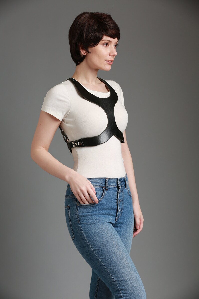 May include: A white t-shirt with a black leather harness. The harness has a unique design with cutouts and straps.