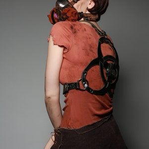 May include: A woman wearing a red shirt with a black leather harness and a gas mask with a floral pattern. The harness has a biohazard symbol design.