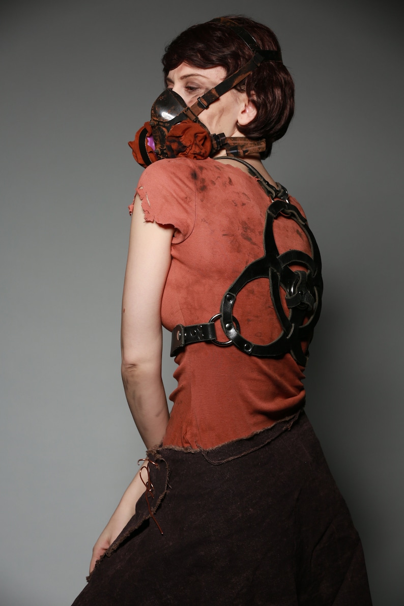 May include: A woman wearing a red shirt with a black leather harness and a gas mask with a floral pattern. The harness has a biohazard symbol design.
