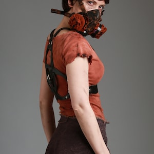 May include: A woman wearing a brown and black gas mask with a red and brown fabric filter. She is wearing a brown, distressed, sleeveless top with a black leather harness. She is also wearing a brown, distressed skirt.