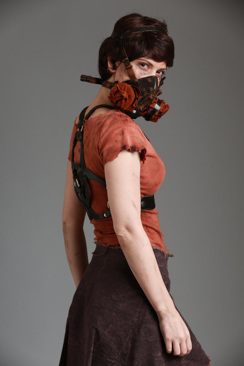 May include: A woman wearing a brown and black gas mask with a red and brown fabric filter. She is wearing a brown, distressed, sleeveless top with a black leather harness. She is also wearing a brown, distressed skirt.