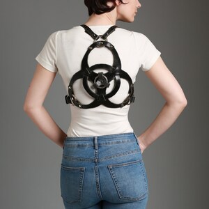May include: A woman wearing a black leather harness with a circular design over a white t-shirt and blue jeans.