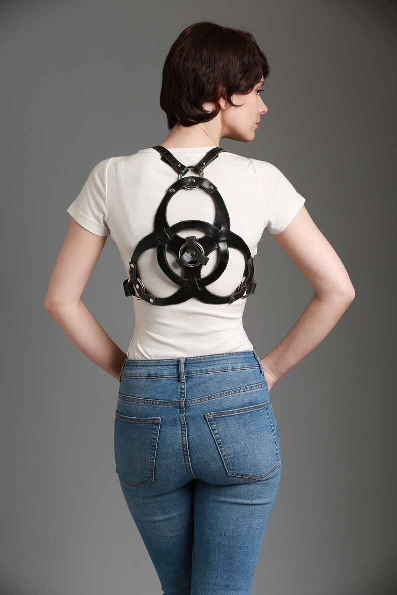 May include: A woman wearing a black leather harness with a circular design over a white t-shirt and blue jeans.