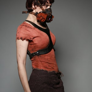 May include: A woman wearing a red shirt with a black leather harness and a brown skirt. She is wearing a gas mask with a red fabric covering the mouthpiece.