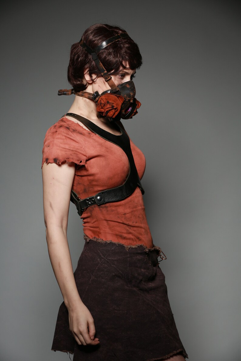 May include: A woman wearing a red shirt with a black leather harness and a brown skirt. She is wearing a gas mask with a red fabric covering the mouthpiece.