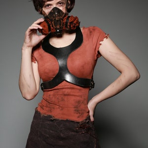 May include: A woman wearing a brown, distressed, short-sleeved shirt with a black leather harness and a brown, distressed skirt. She is wearing a gas mask with orange and brown accents.