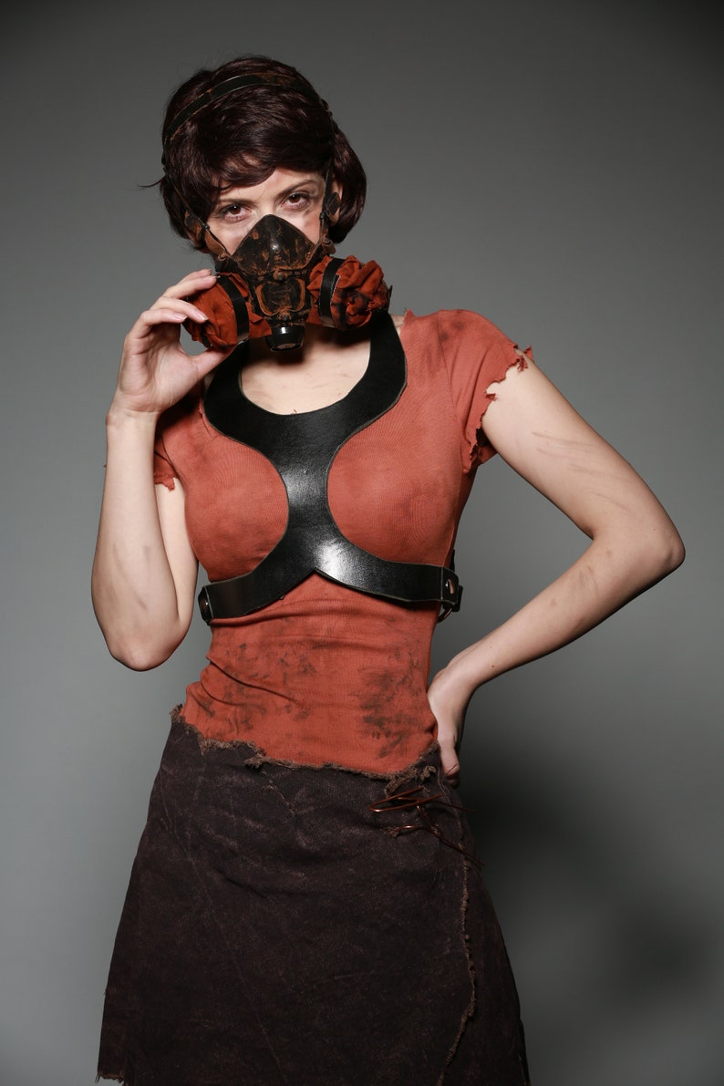 May include: A woman wearing a brown, distressed, short-sleeved shirt with a black leather harness and a brown, distressed skirt. She is wearing a gas mask with orange and brown accents.