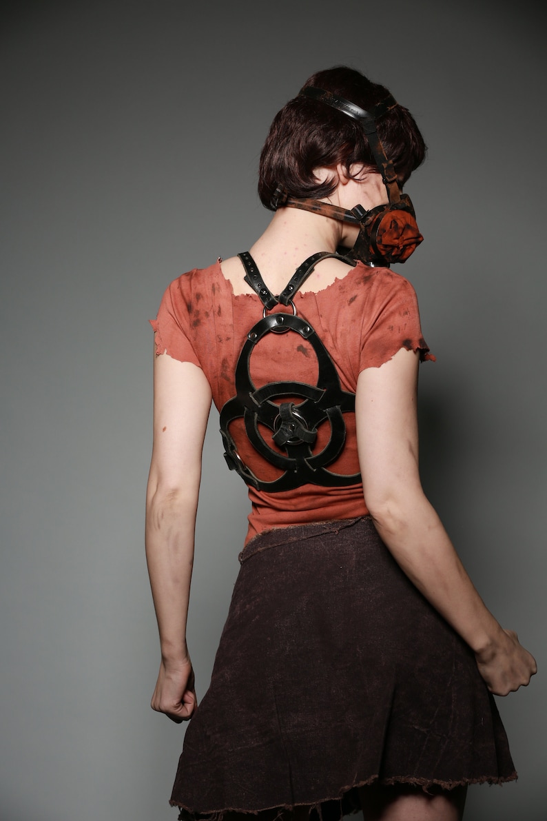 May include: A woman wearing a red shirt with a black leather harness featuring a biohazard symbol. She is also wearing a brown skirt and a gas mask.