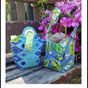 May include: Two reversible baby bindle bags, one with a blue and green geometric pattern and the other with a blue and green floral pattern. The bags are 4 inches by 4 inches by 10 inches and are made with fat quarters. The bags are sitting on a wooden bench. The text "Sisters' Common Thread Baby Bindle" is at the top of the image. The text "FULLY REVERSIBLE - FAT QUARTER FRIENDLY" is below the bags. The text "Size: 4"X4"X10"" is below that. The text "Full-size pattern piece included" is below that. The text "www.sisterscommonthread.com" is below that. The text "Designed by Yvonne Cabella" is below that. The text "Item # SCT10164" is below that.