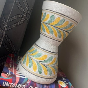 May include: A white ceramic vase with a textured surface and a design of yellow and green leaves. The vase has a wide mouth and a narrow base.