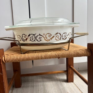 May include: White oval casserole dish with a gold and brown floral design and a glass lid. The dish is sitting on a wooden stand with a woven straw seat.