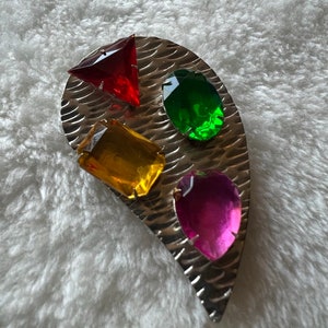 May include: A silver brooch with four colorful glass stones. The stones are red, green, yellow, and pink. The brooch is shaped like a teardrop.