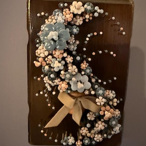 May include: A decorative wall hanging featuring a floral arrangement. The arrangement includes blue, white, and peach-colored flowers and small pearl-like accents. A tan ribbon is tied at the base of the arrangement, set against a dark brown background.
