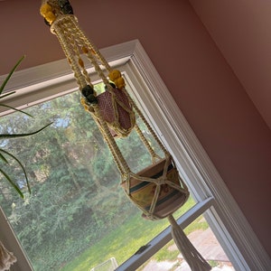 May include: A macrame plant hanger with yellow and green beads hangs from a hook in a room with a window. The hanger has two pots, one with a pink and white pattern and the other with a brown and green pattern.