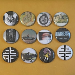 May include: A collection of twelve round pinback buttons with various designs. The buttons feature images of a cat, a compass, a person, and text that reads "JAWBREAKER Dear You" and "LIVE 4/30". The buttons are on a yellow background.