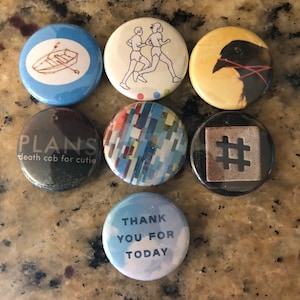 May include: Seven round buttons with various designs. The buttons are blue, brown, black, white, and yellow. The designs include a boat, two people running, a bird, a hashtag, a geometric pattern, and the words "Plans death cab for cutie" and "Thank you for today".