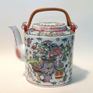 May include: A white ceramic teapot with a floral design and a woven handle. The teapot features a detailed pattern of flowers in pink, blue, yellow, and green. The handle is made of woven brown material, and the spout has a red sunburst design.