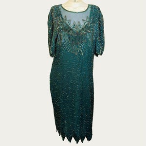 May include: Emerald green beaded dress with short sleeves and a sheer neckline. The dress features intricate beadwork and a scalloped hemline. The dress is a knee-length style, perfect for a special occasion.